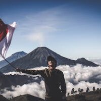 Man Wearing Black Crew-neck Sweater Holding White and Red Flag Standing Near Mountain Under Blue and White Sky